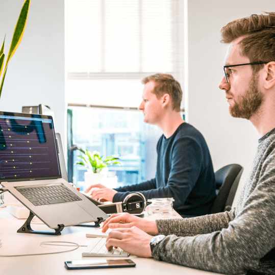 Two men sit at a bright shared desk, working on laptops with headphones and a plant nearby.